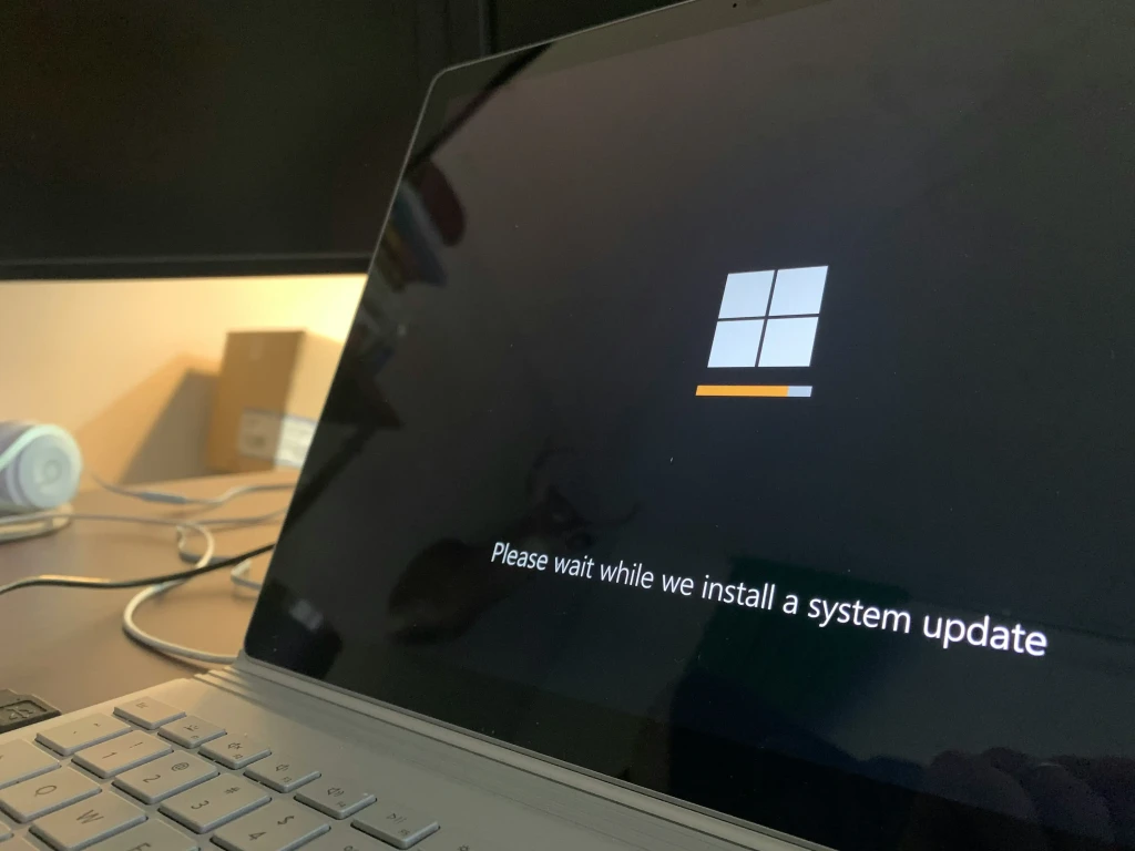 Bærbar computer på et bord viser Windows-logo og systemopdatering-skærm. Baggrund har kabler og en papæske.