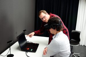 Auf dem Bild sieht man Dennis mit einer Virtual Reality Brille auf dem Kopf. Thorsten zeigt ihm etwas auf dem Laptop obwohl Dennis nichts sehen kann.