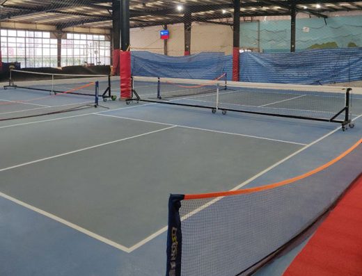 Indoor view of two sky blue pickleball courts at Asoke Sports Club in Bangkok, Thailand. The light blue playing surfaces span the visible foreground with thick white nets and posts dividing the spaces. Additional volleyball courts are partially visible further back beyond fencing.