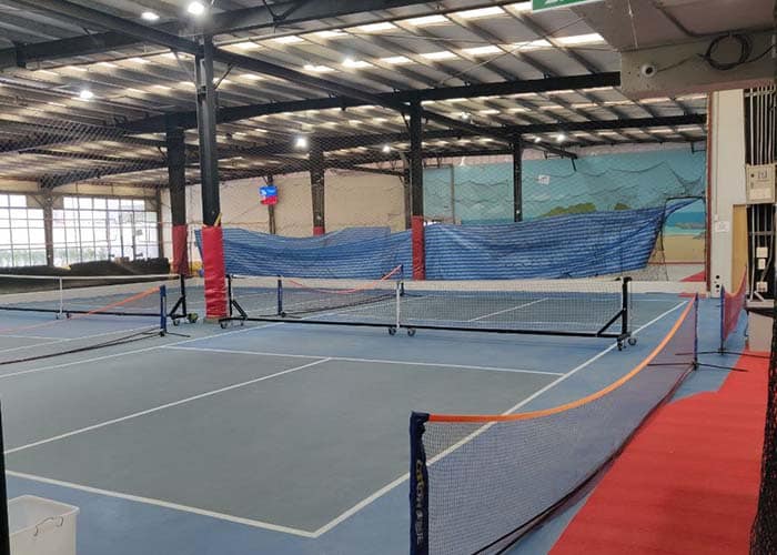 Indoor view of two sky blue pickleball courts at Asoke Sports Club in Bangkok, Thailand. The light blue playing surfaces span the visible foreground with thick white nets and posts dividing the spaces. Additional volleyball courts are partially visible further back beyond fencing.