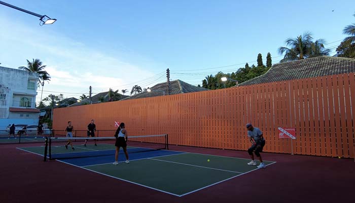 Players engaging in a pickleball match at Shark Bite court, Phuket, with surrounding buildings and tall palms at sunset.