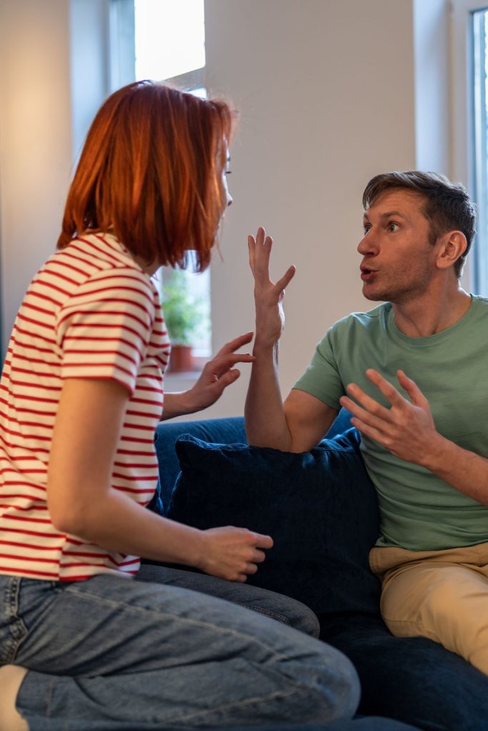 Woman is crouched on the couch talking to man, while he is responding to her with his hands. 