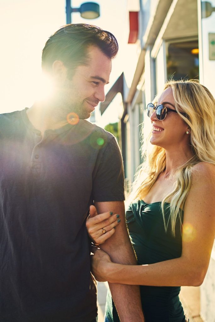 A woman is holding unto the arm of a man closely. She and him are smiling and looking at each other's eyes