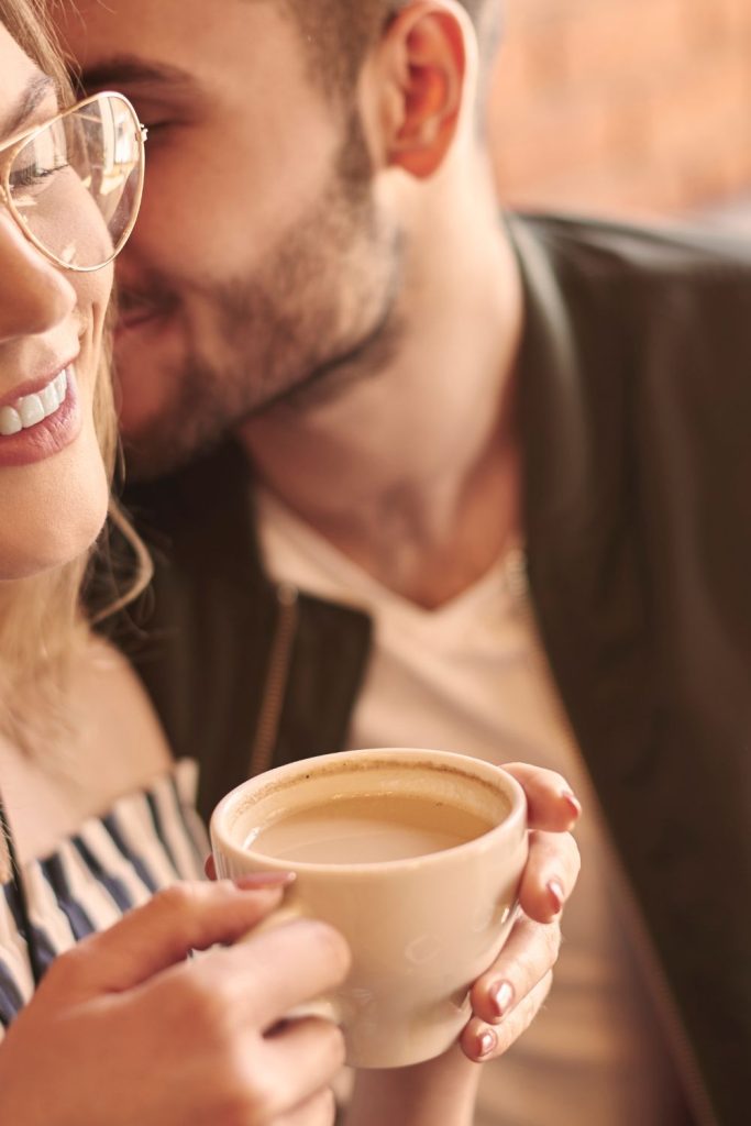 Man with his lips close to woman's ear. She listens intently while holding a coffee cup