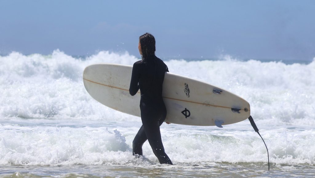 Girl wearing a wetsuit and carrying a surfboard, walking into the water