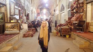 Ingrid Sørensen, dancer and painter, stands in the middle of market Bazaar in Isfahan, Iran wearing a yellow hijab loose around her face, and a blue sweater and dark jeans. Image is slightly washed out and dusty.