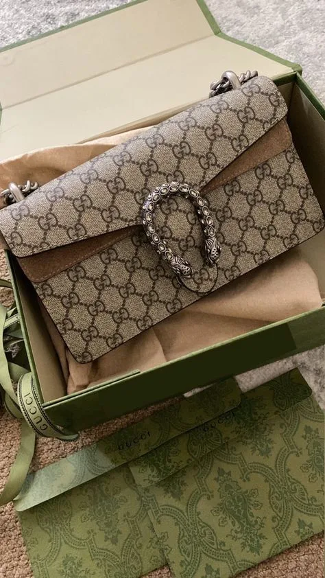 An open box on a carpeted floor displays a Gucci Dionysus bag, presenting its monogram canvas and the detailed silver chain link clasp. The green and gold packaging of the box adds a luxurious feel to the presentation.