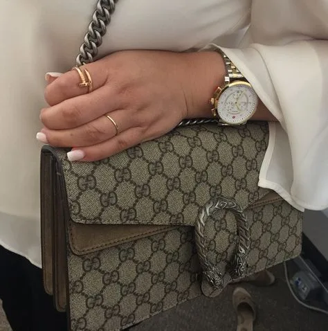 A close-up view of a hand elegantly adorned with multiple gold rings and a gold-toned wristwatch, clutching the handle of a Gucci Dionysus bag. The bag features the iconic Gucci monogram and a unique tiger head spur closure.