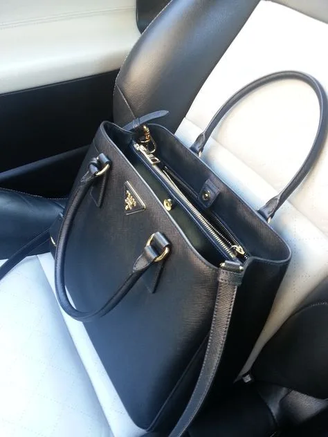 An open black Prada Saffiano tote bag rests on a white leather car seat, revealing a spacious interior with pockets, perfect for organization. The bag's structured silhouette and gold-tone hardware exude a sense of classic style and functionality.