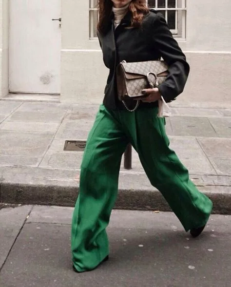 A person strides forward on a city sidewalk, donning a sleek black blazer paired with striking green trousers. They hold a Gucci Dionysus bag with a distinctive chain strap and a textured, quilted pattern.