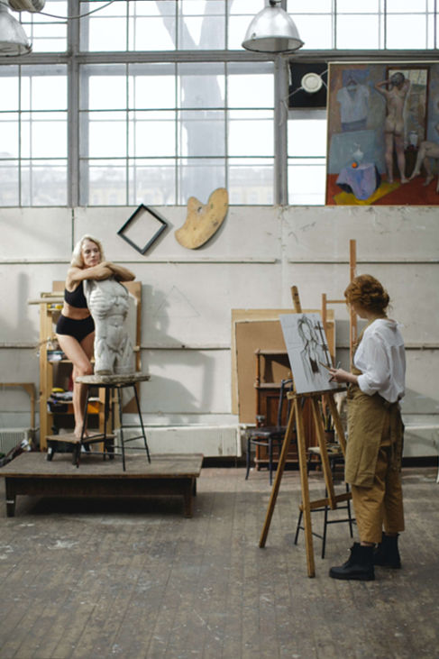 Two artists working together in an art studio, one painting on a canvas while the other is modeling for a sculpture