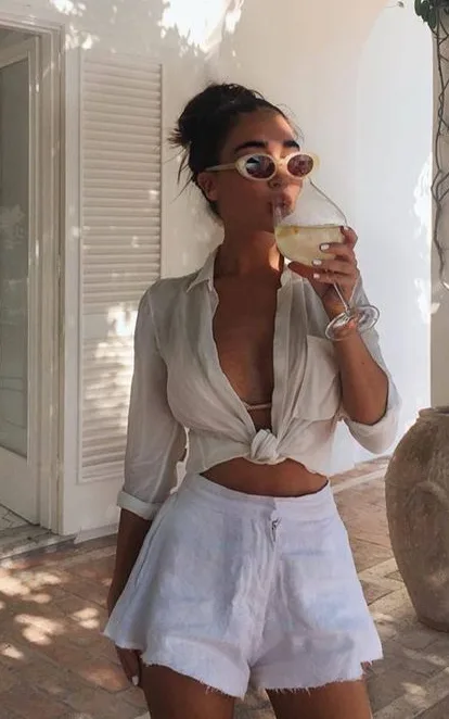 Woman with sunglasses drinking a glass of wine in a white vacation outfit. 