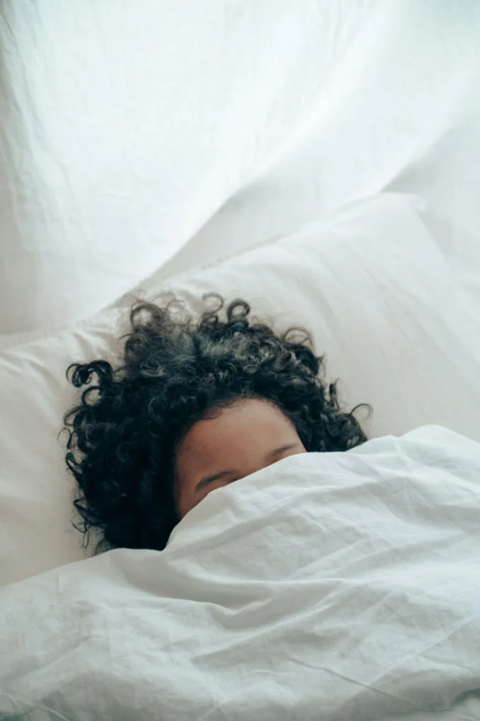 Woman optimizing her sleep in a light, bright and white room sleeping under the covers with only her hair showing.