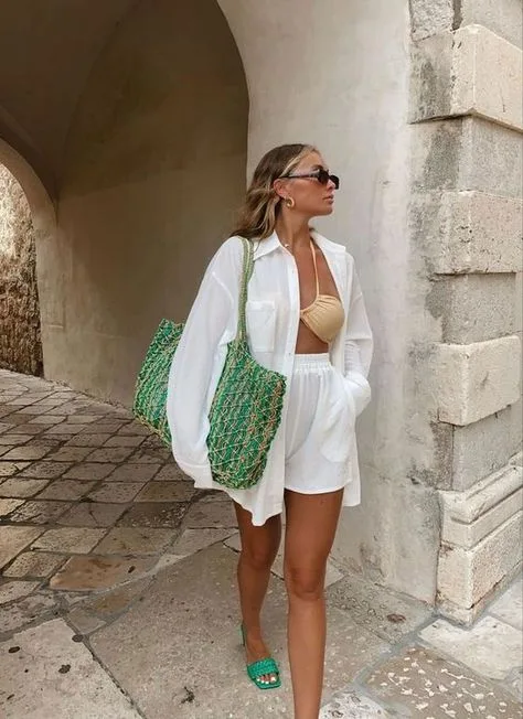 Bohemian-inspired vacation look with a woman in a white shirt dress and bikini top, accessorized with a green tote and sunglasses, standing in a historic cobblestone alleyway.