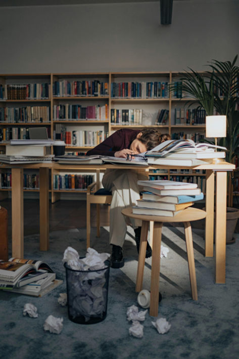 a woman is tired and resting her head on a table covered in lots of books and paper
