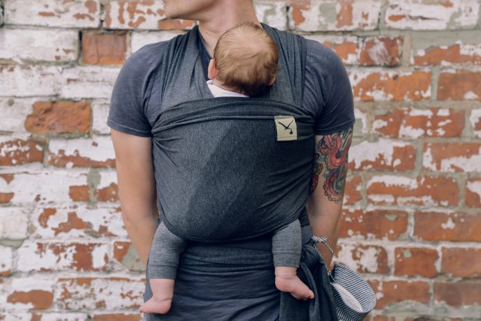 Person shown from neck down carrying baby in wrap-style baby carrier with baby facing inwards.