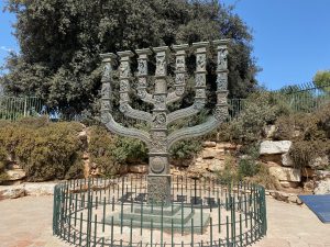 Utendørs jødisk menorah med åtte lysestaker og en sjette arm, omgitt av gjerde, i en park med trær o.