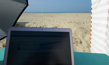 Een laptop met op de achtergrond de zee, een strandzeil en een parasol.