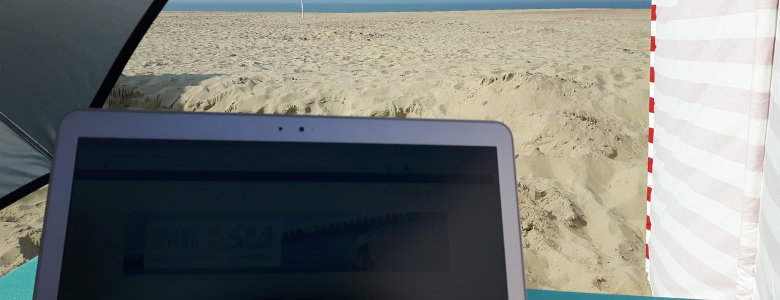 Een laptop met op de achtergrond de zee, een strandzeil en een parasol.