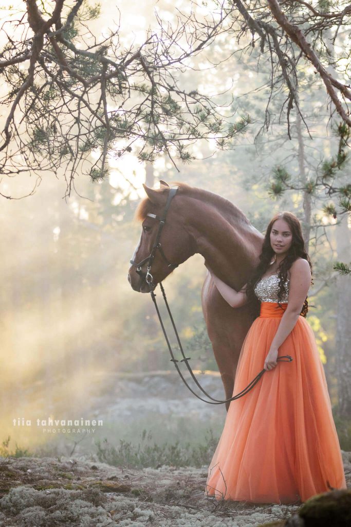 Suomenhevosruuna Usvasuon Donjuan oranssiin prinsessamekkoon pukeutuneen omistajansa kanssa kallion laella auringonsäteiden siivilöityessä usvan ja puiden läpi