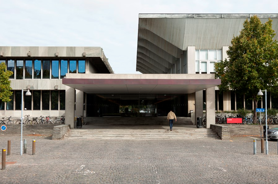 Et eksteriørbillede af en bygning tilknyttet Polyteknisk Kollegieselskab. Bygningen har en moderne arkitektur med betonfacader og store vinduer, der lader naturligt lys strømme ind. Foran indgangen er der brede trappetrin og et overhængende tag, der danner en overdækket indgangsportal. Området omkring bygningen er brolagt og har flere cykelstativer, som er fyldt med cykler, hvilket afspejler den studerendes livsstil. En person går mod indgangen, hvilket tilføjer en dynamisk dimension til billedet. Polyteknisk Kollegieselskab tilbyder moderne faciliteter og en praktisk beliggenhed for studerende, der er en del af det tekniske universitetsmiljø.