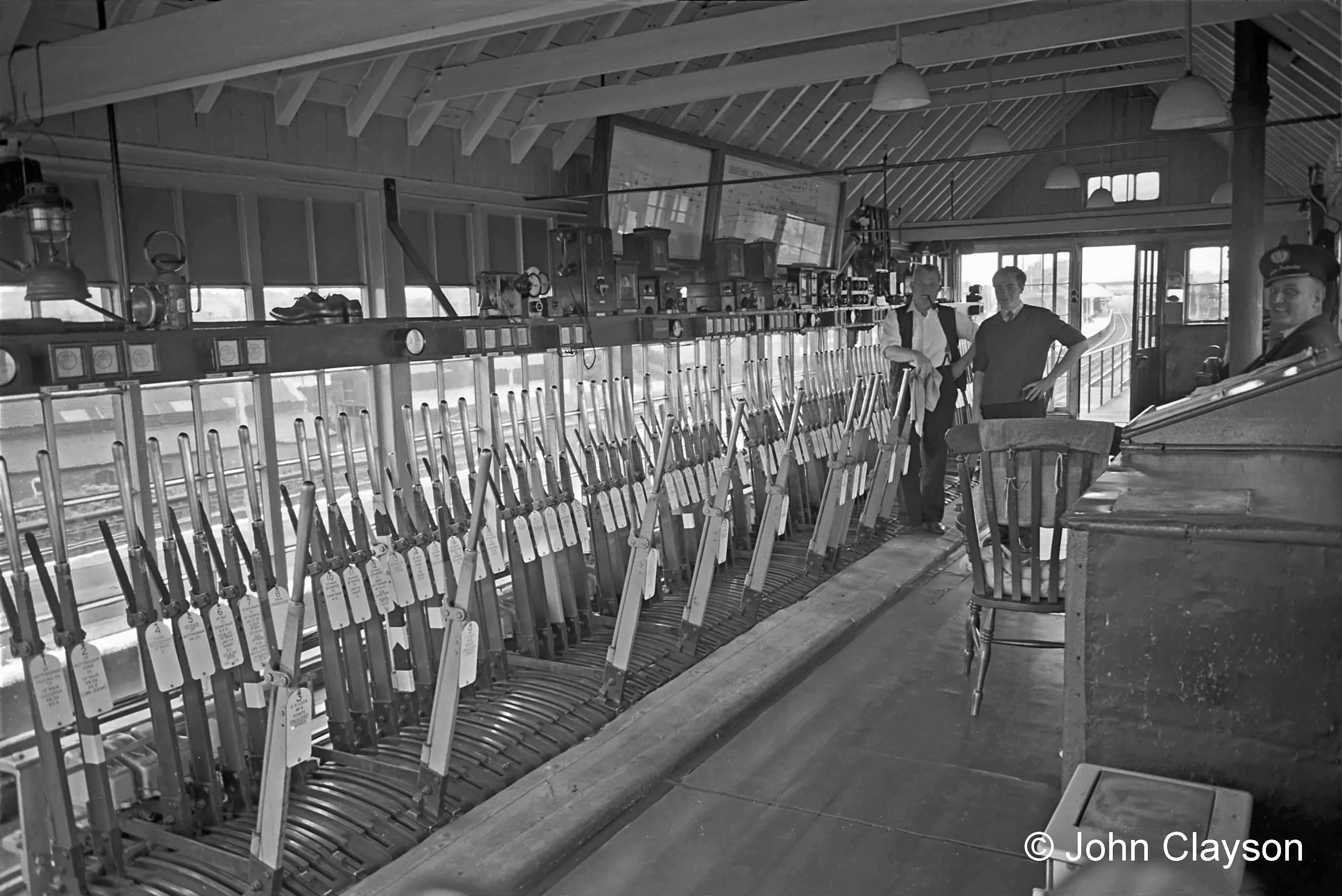 On duty were Signalman Albert Eldridge - with pipe and trademark cloth - and his Telegraph Lad Dennis Gray. The view through the windows at the far end is south, through the main line platforms. Inside the box everything is sparkling clean and tidy, as is customary. At top left a ‘Tilley’ paraffin vapour lamp hangs, ready to illuminate the box in case of power failure. On the block shelf just below are neatly rolled flags and, next to them, a signalling lamp for hand signalling by day and by night respectively. Further along is a pair of well-worn, brightly polished shoes! No doubt they belong to a man on another shift who, on arrival, would change into them from his outside footwear to avoid scuffing that highly polished lino. Photograph by Cedric A. Clayson.