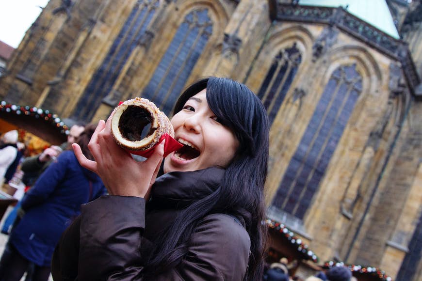 Porträtt av en leende ung asiatisk kvinna i en vinterrock som äter en trdelník (en rund bakelse) i Prag framför en kyrka