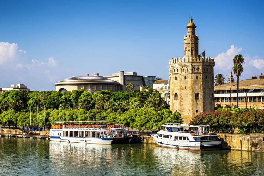 Sevillas Torre del Oro, ett cirkulärt stentorn med en minaret intill floden;  vita båtar ligger förtöjda framför den och det finns träd och buskar på vardera sidan om tornet.