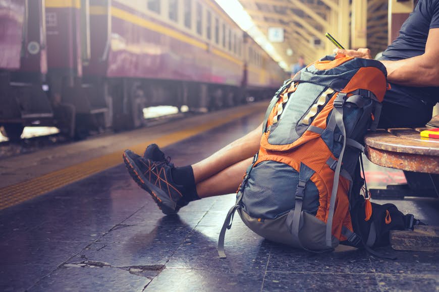 En man sitter på en bänk på en tågstation och tittar på sin telefon. Det ligger en stor ryggsäck på golvet bredvid honom, och ett lila tåg står stilla vid perrongen.
