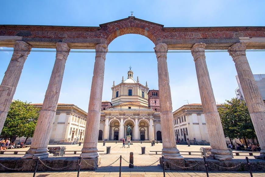 San Lorenzo-kolonnerna, en rad av stenpelare från romersk tid med en trädkantad piazza och en kyrka bortom.