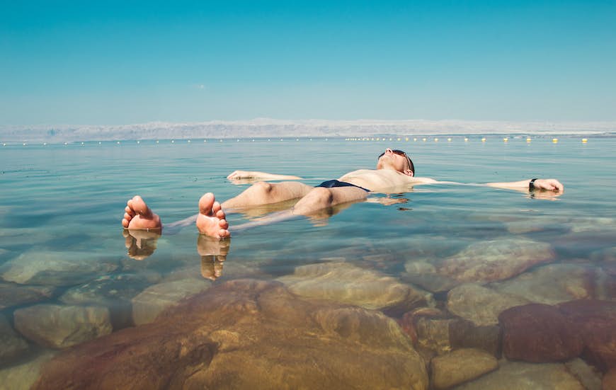 En man som bär solglasögon och en mörk Speedo flyter på ryggen i det salta blå vattnet i Döda havet, Israel