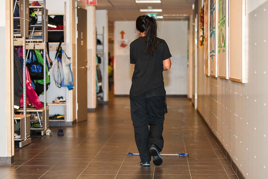 A woman, seen from behind, is sweeping and cleaning the floors in a school corridor.