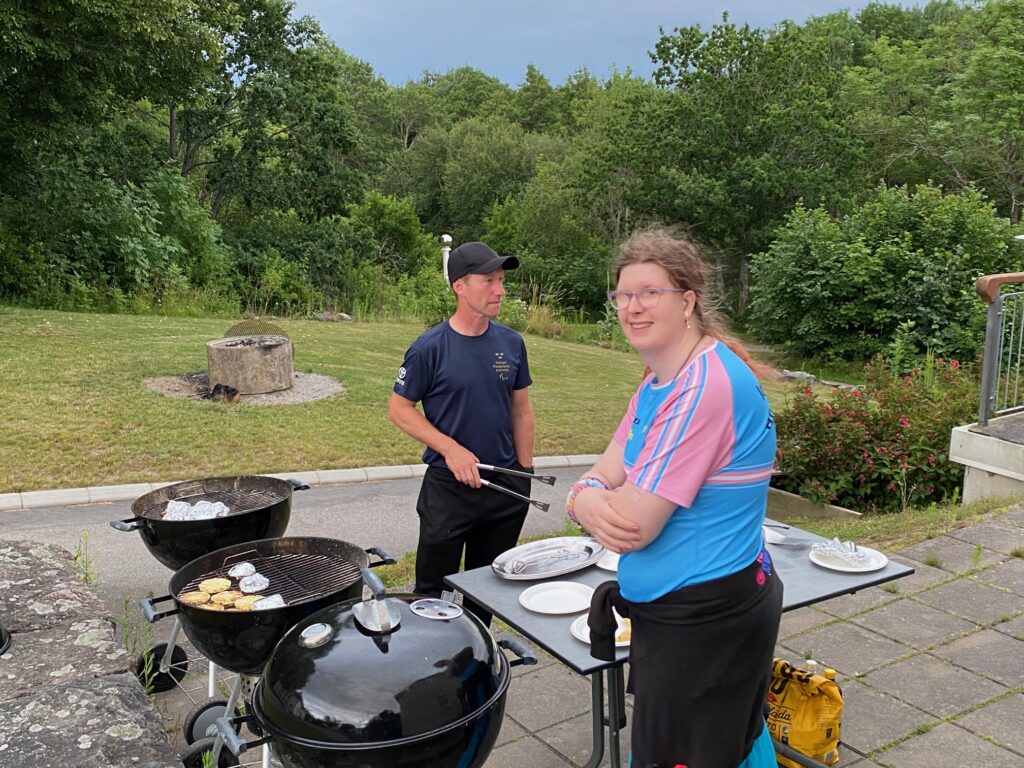 Två ungdomar står utomhus i en trägård, bredvid tre grillar med mat på. En håller i en grilltång.
