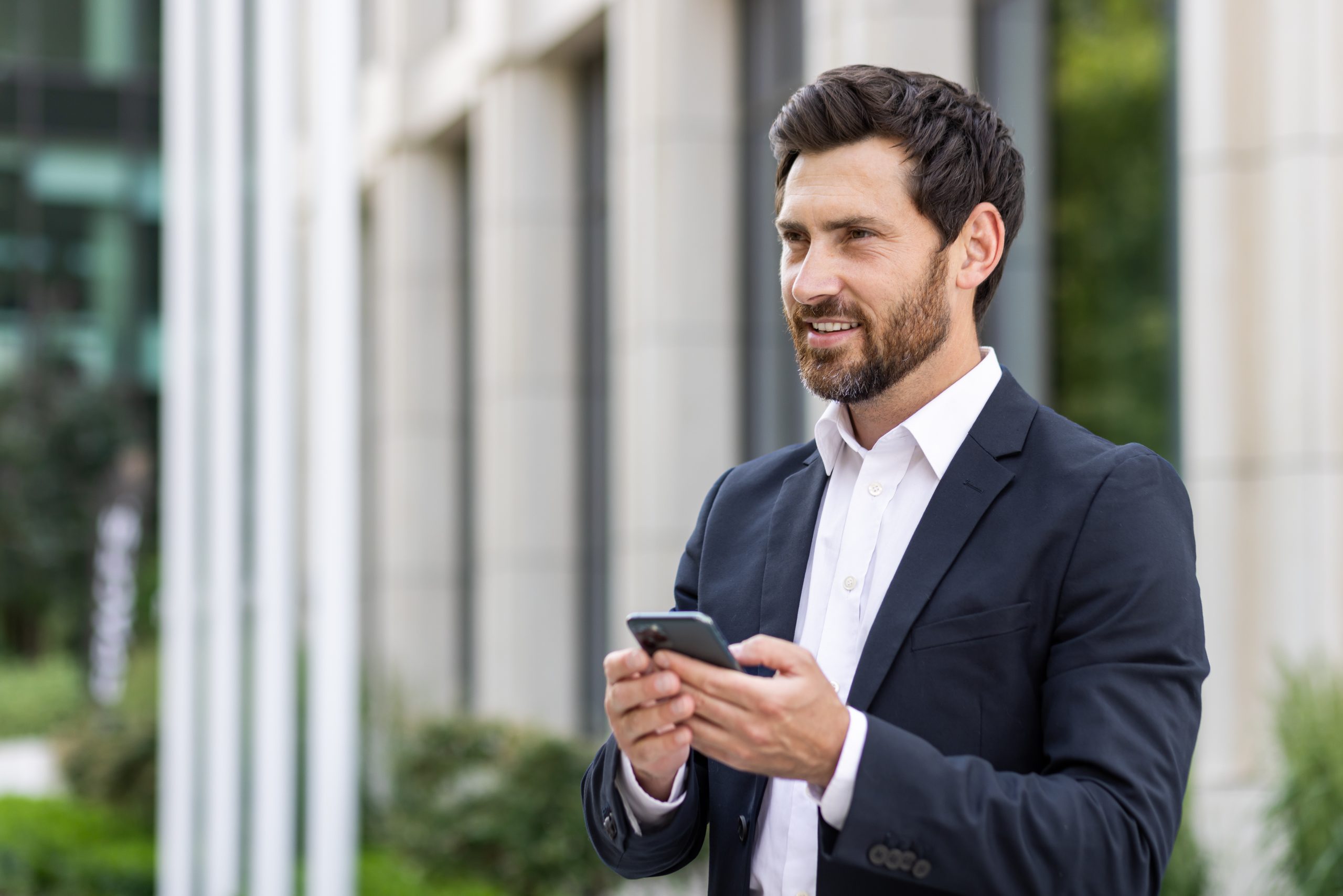 Close-up photo of a young businessman standing in a business suit and using a mobile phone, smiling and looking to the side