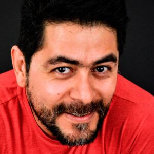 A man smiling, with black hair, beard and moustache. Wearing a red t-shirt.