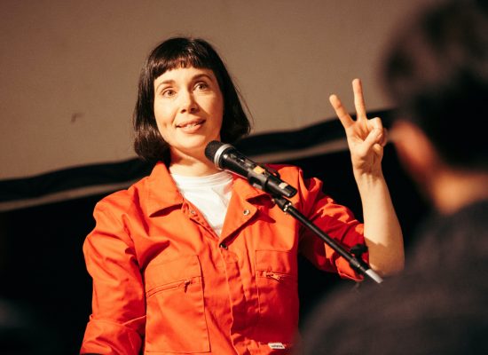 A white woman with dark hair, wearing a red jumpsuit, is smiling and making a V for Victory sign with her left hand.