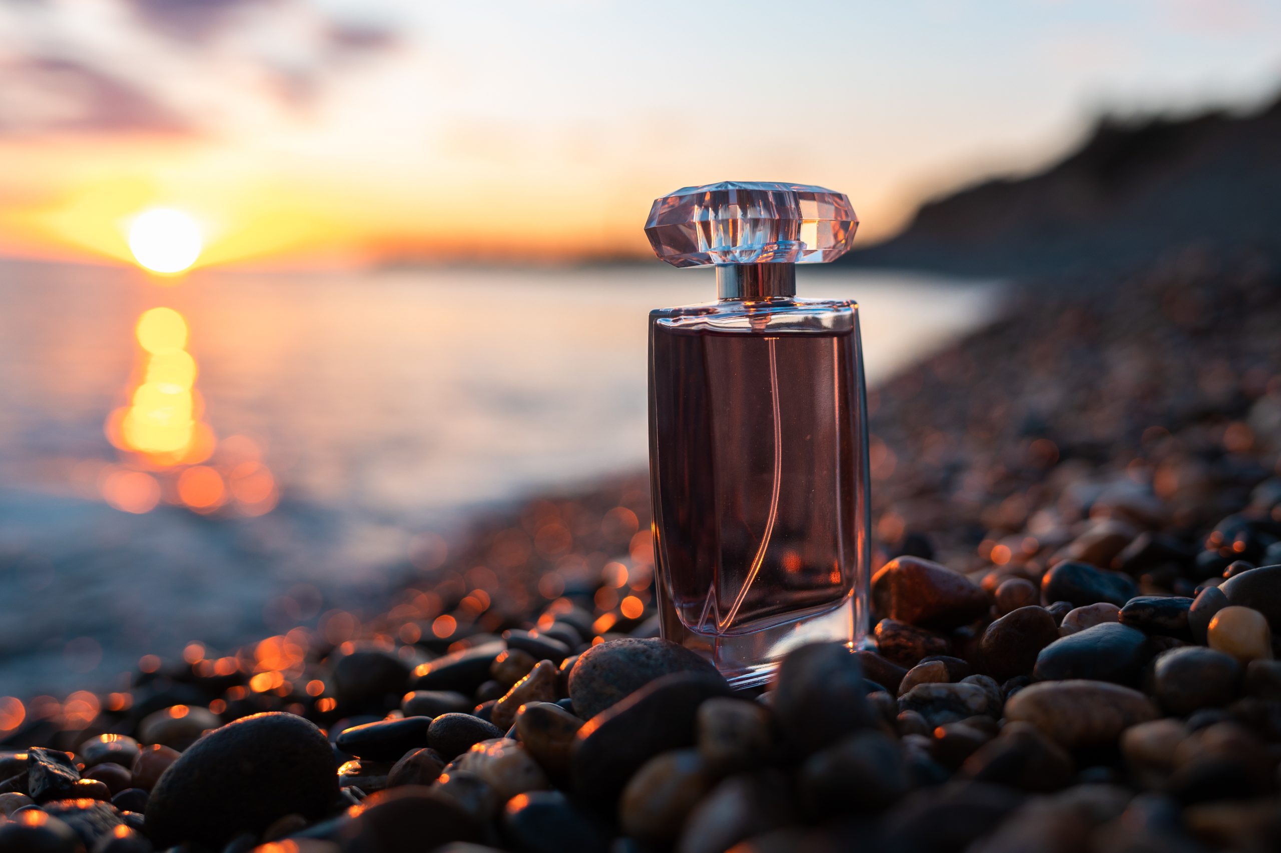 Perfumery and marketing. Lilac glass perfume bottle on a pebble beach, on the seashore, close-up. Sunset on the background. Luxury Perfume presentation concept