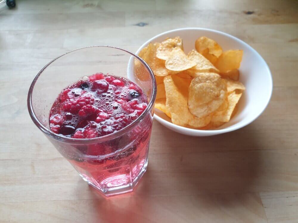 Ein Glas Wildberry von Schweppes mit gefrorenen Waldbeeren und eine kleine Schale Kartoffelchips.