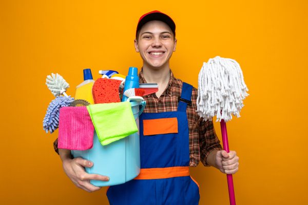 smiling-young-cleaning-guy-wearing-uniform-cap-holding-bucket-cleaning-tools-with-mop
