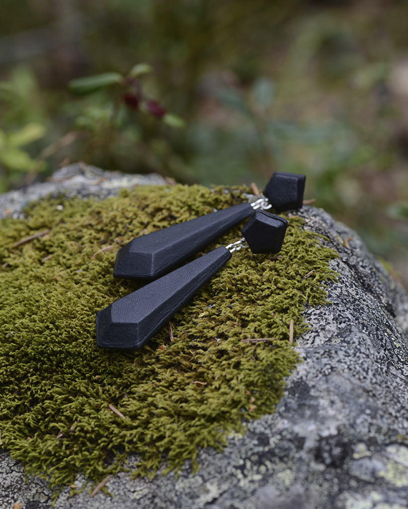 Side view of a pair of black leather and silver earrings on a grey stone in a forest setting. The earrings are drop shaped with faceted edges.