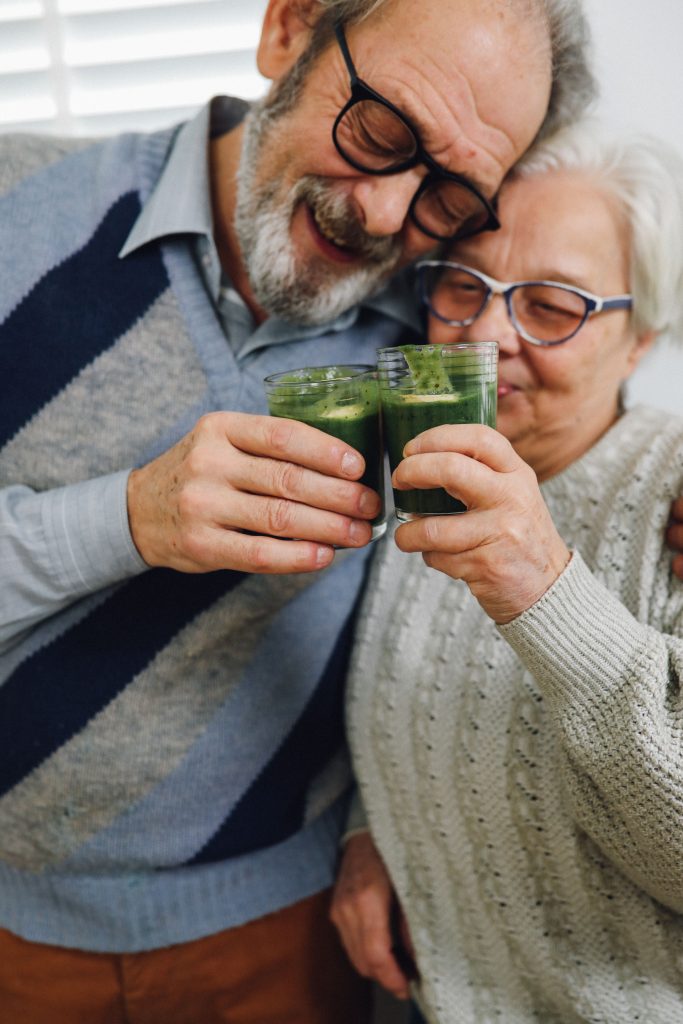 Happy senior couple clinking glasses of green smoothies, embracing moments of joy indoors.