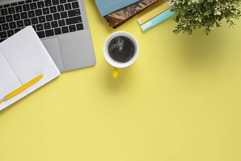 planung Auf einem gelben Tisch steht ein Laptop, eine Kaffeetasse, eine Pflanze und ein paar Stifte mit Notizblöcken