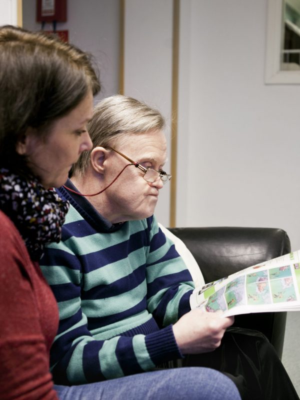 Caregiver looking at mentally challenged senior man reading magazine at home