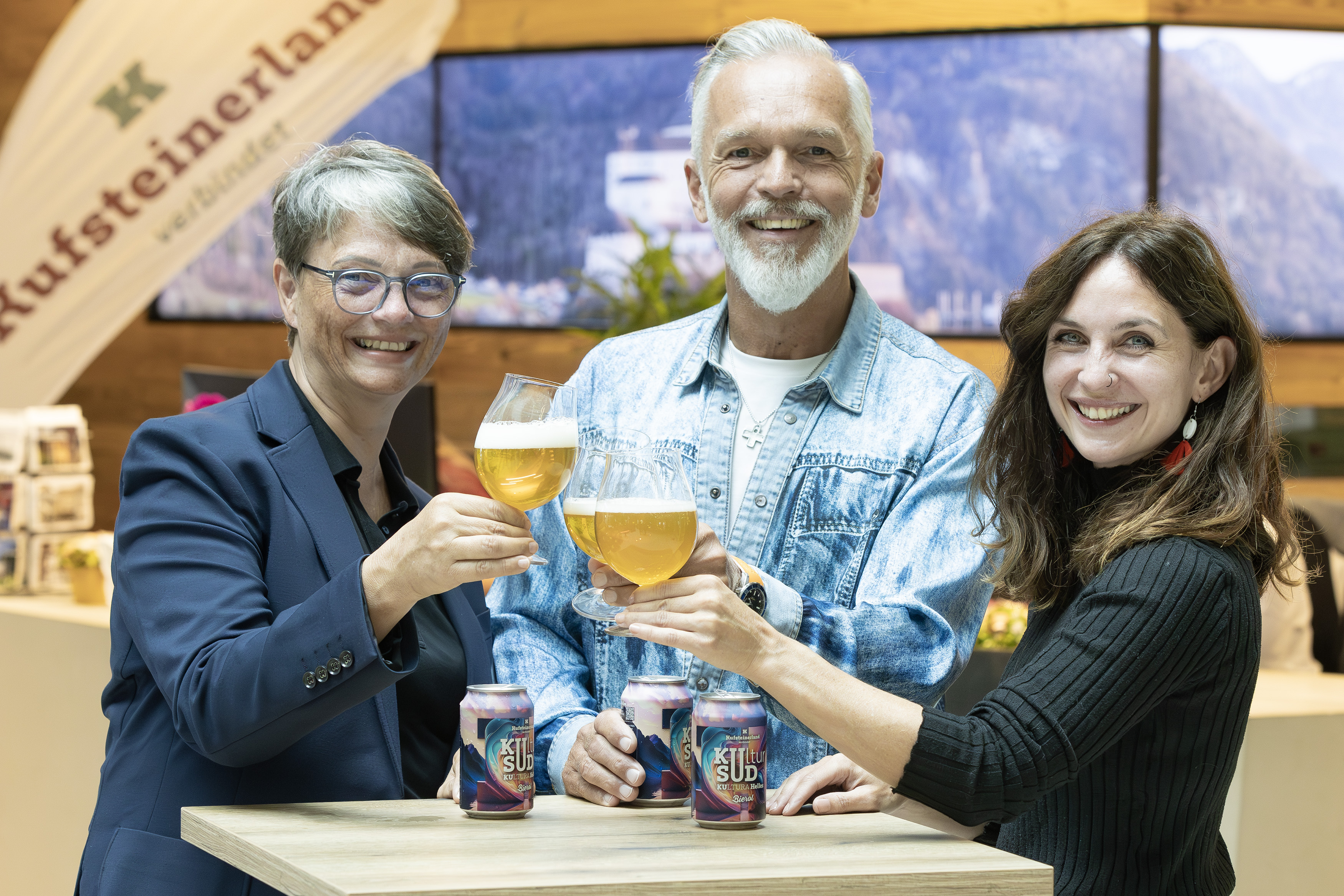 Drei Personen lächeln und stoßen mit Biergläsern an, während sie vor einem Stand mit einem „Kufsteinerland“-Banner stehen. Auf dem Tisch vor ihnen stehen mehrere Dosen des Bieres „Kultur Sud“. Die Atmosphäre ist freundlich und einladend. Die Personen sind von links Sabine Mair, Georg Hörhager (beide vom TVB Kufsteinerland) und Lisa Luginger-Bichler von Bierol