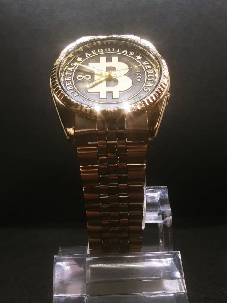 The dial is styled like circulated coin the raised areas are gold coloured with a brushed finish and the lower pressed arease are black with a light brushing of gold colour. The dial reads, Libertas,  Aequitas, Veritas, In cryptography we trust dated 2009 when bitcoin was born