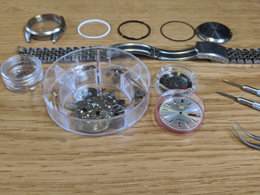 Wellingtime UK Watch Repair service - Watch Strip Down, all components parts after they have been cleaned for watch repair. The dial, day/date wheel, balance wheel, case, dial spacer, movement holder, gasket caseback gasket and jubilee bracelet can all be seen individually spaced out in the centre there is a parts box which contains all of the watch movement component parts for repair. All work undertaken in the UK.