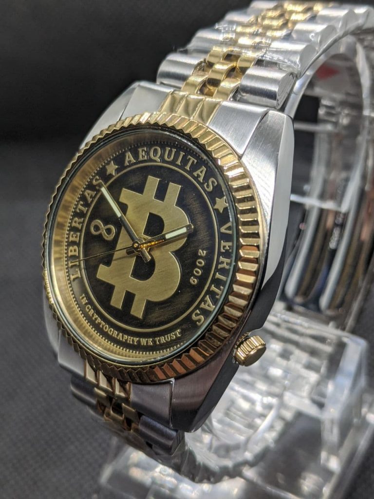 The dial is styled like circulated coin the raised areas are gold coloured with a brushed finish and the lower pressed arease are black with a light brushing of gold colour. The dial reads, Libertas,  Aequitas, Veritas, In cryptography we trust dated 2009 when bitcoin was born