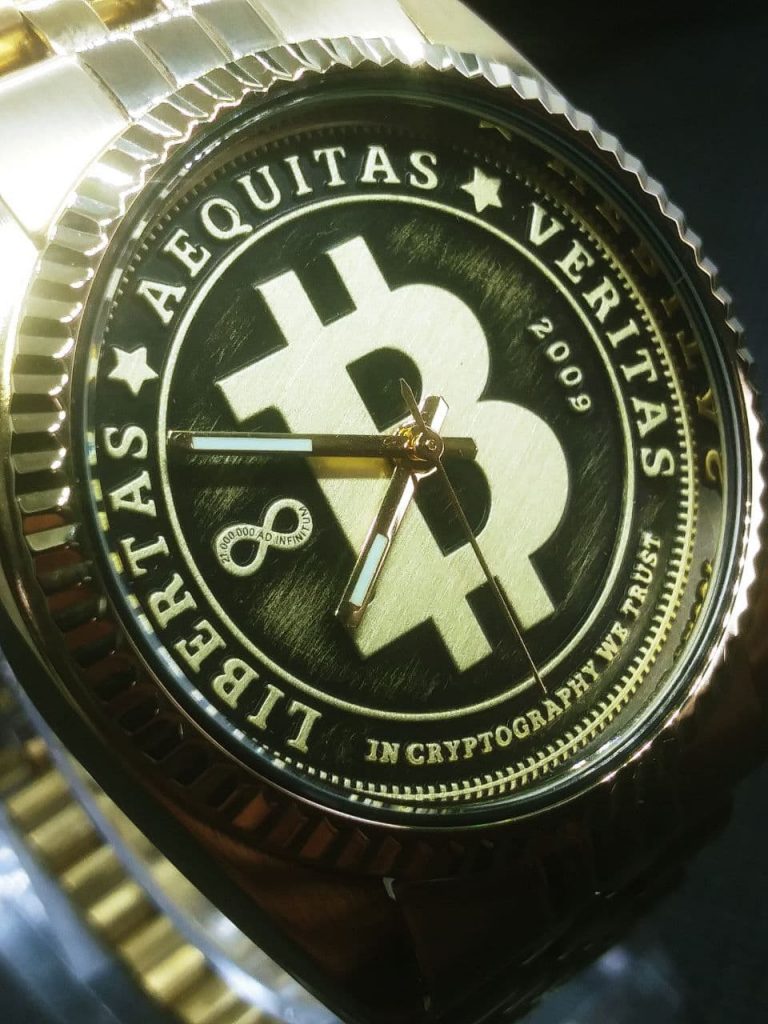 The dial is styled like circulated coin the raised areas are gold coloured with a brushed finish and the lower pressed arease are black with a light brushing of gold colour. The dial reads, Libertas,  Aequitas, Veritas, In cryptography we trust dated 2009 when bitcoin was born