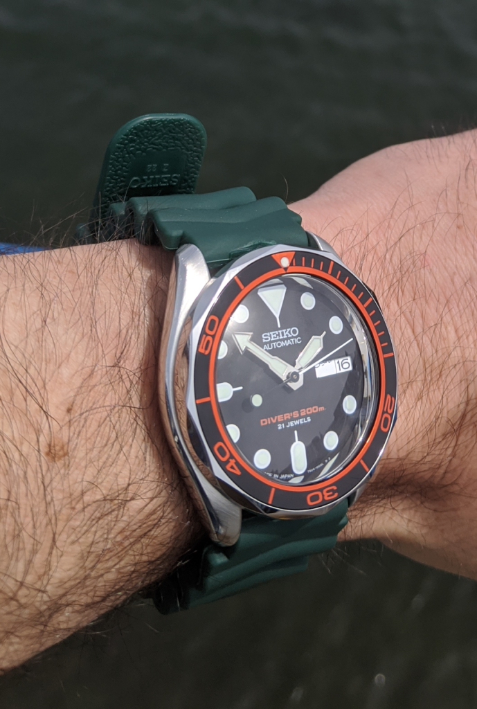 Seiko SKX007J Seamaster Mod - Black Orange insert set in a seamaster style bezel, single dome insert  no beveled edge. Displated on the wrist againts the green water of the harbour falmouth - watch repair cornwall