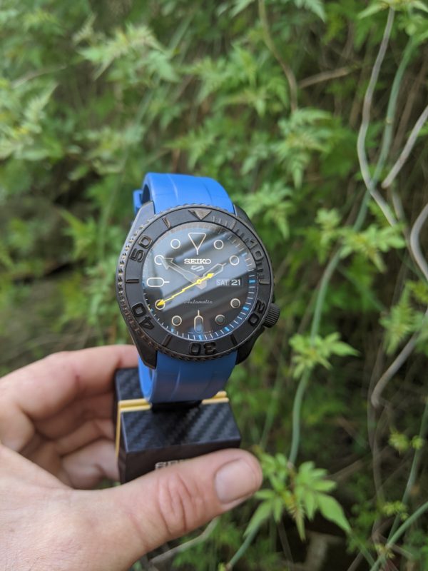 Seiko SRPD79 Blackout Mod - Blue Monstraps 2 peice. YM insert, chapter ring with blue hour markers, yellow lighting second hand, double dome sapphire with blue AR coating - displayed on a carbon seiko display stand infront of foliage